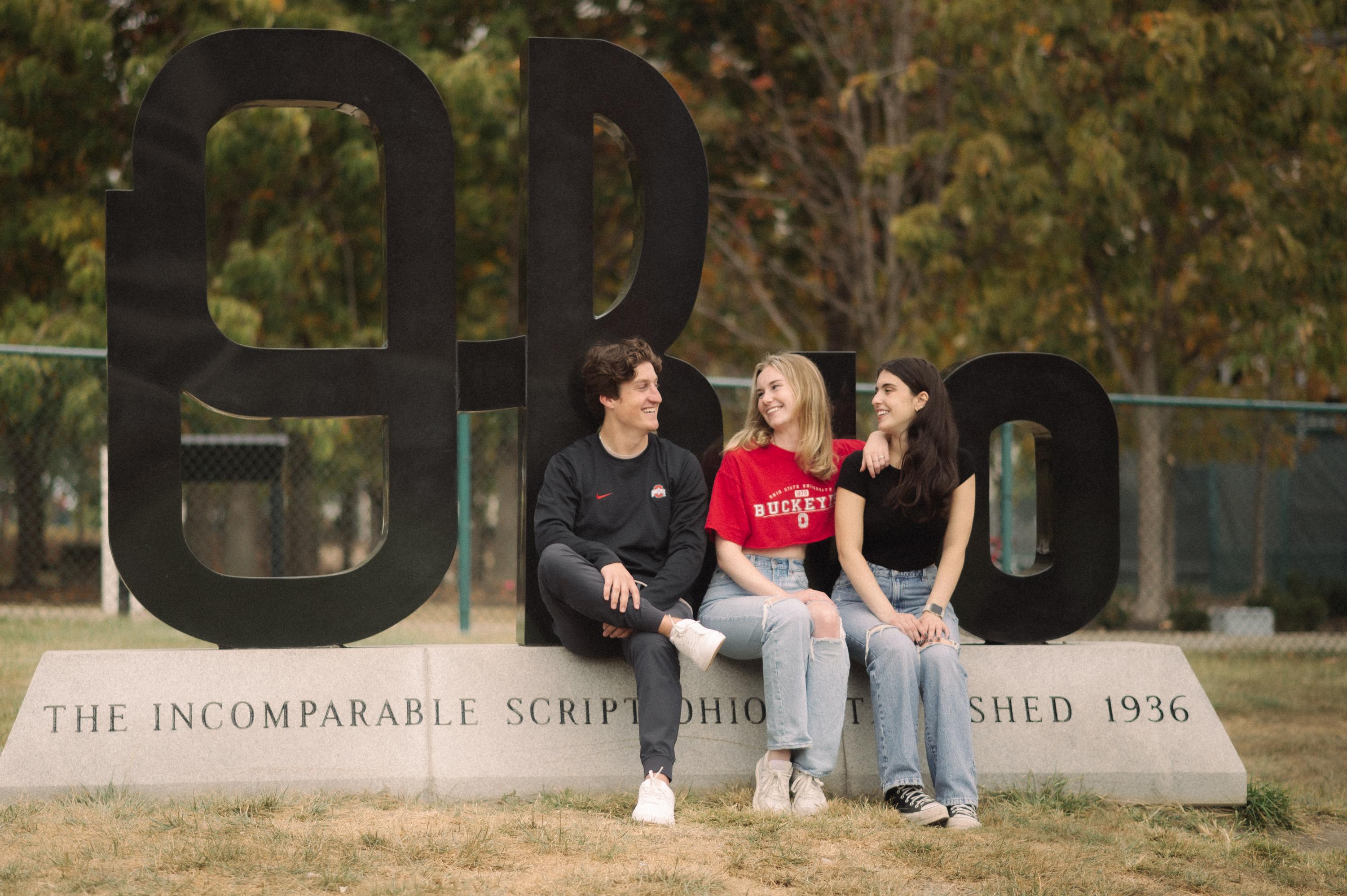 three OSU students sitting on camous near the Ohio sign